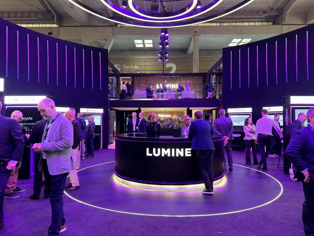 A half moon desk at the center of a large convention booth with people around and Lumine on the front of the desk